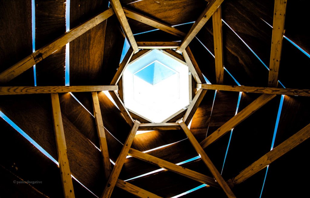 A sky-facing photo taken from the inside of a geometric art piece, sunlight shines through a hexagonal portal at image centre, surrounded by a complimentary wooden structure