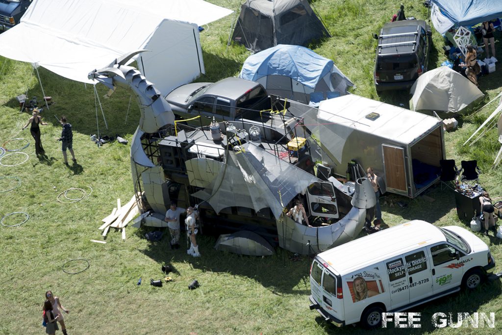 A vehicle that has been converted in to a dragon (known as Heavy Meta) sits in a grassy field during the day, surrounded by vans, trailers and tents.